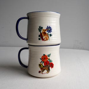 K&ouml;nnte beinhalten: Zwei cremefarbene Keramikbecher, die &uuml;bereinander gestapelt sind, mit blauen Griffen und R&auml;ndern. Jeder Becher hat eine andere Illustration: einer mit einem Kaninchen und blauen Blumen, der andere mit einer Katze und roten Blumen. Die Becher haben eine gerippte Textur.