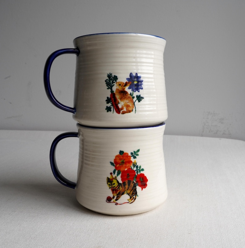 K&ouml;nnte beinhalten: Zwei cremefarbene Keramikbecher, die &uuml;bereinander gestapelt sind, mit blauen Griffen und R&auml;ndern. Jeder Becher hat eine andere Illustration: einer mit einem Kaninchen und blauen Blumen, der andere mit einer Katze und roten Blumen. Die Becher haben eine gerippte Textur.
