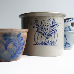 May include: A collection of stoneware pottery with blue designs. The largest pot is light brown with a blue floral motif. Other pots include a light brown pot with blue leaves and a stack of smaller blue and white pots.