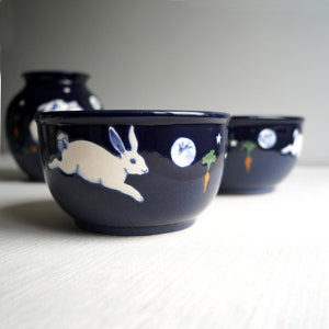 May include: A set of three blue ceramic bowls with white rabbit and moon designs. The bowls are hand-painted with a whimsical style.