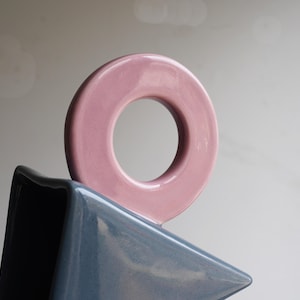 May include: A contemporary ceramic sculpture. A light pink, circular form sits above a triangular, blue-grey shape. The glossy ceramic finish reflects light, emphasizing the smooth surfaces and geometric design.
