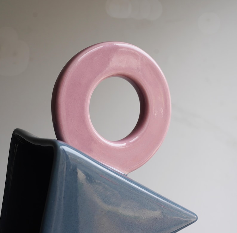 May include: A contemporary ceramic sculpture. A light pink, circular form sits above a triangular, blue-grey shape. The glossy ceramic finish reflects light, emphasizing the smooth surfaces and geometric design.