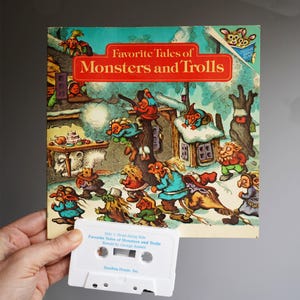 May include: A vintage children's book titled "Favorite Tales of Monsters and Trolls" with a colorful illustration of trolls and monsters in a snowy landscape. A white cassette tape is held below the book, with text that reads "Side 1 Read-Along Side".