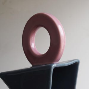 May include: A contemporary ceramic vase featuring a circular handle in a pale pink shade. The base of the vase is a contrasting dark blue-grey colour. The vase has a distinctive, geometric design.
