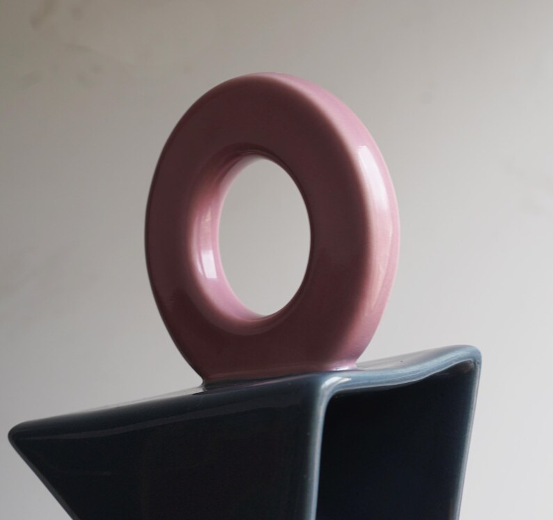 May include: A contemporary ceramic vase featuring a circular handle in a pale pink shade. The base of the vase is a contrasting dark blue-grey colour. The vase has a distinctive, geometric design.