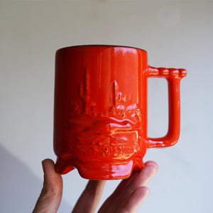 May include: A vibrant, glossy red ceramic mug with a unique handle design. The mug features a raised relief design of a desert landscape with cacti and the text "ROAD RUNNER". The mug is held by a hand.