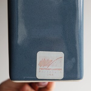 May include: A close-up of a blue-grey ceramic vase with a rectangular shape. The vase has a textured surface. A sticker on the bottom reads "FELTMAN LANGER Porcelain USA".