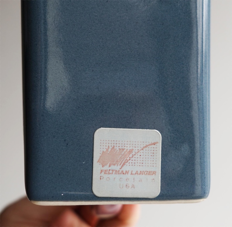 May include: A close-up of a blue-grey ceramic vase with a rectangular shape. The vase has a textured surface. A sticker on the bottom reads "FELTMAN LANGER Porcelain USA".