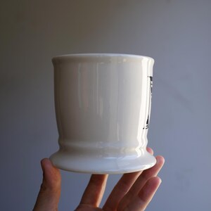 Pu&ograve; includere: Tazza in ceramica bianca con un design bianco e nero. La tazza ha una base larga e un corpo arrotondato.