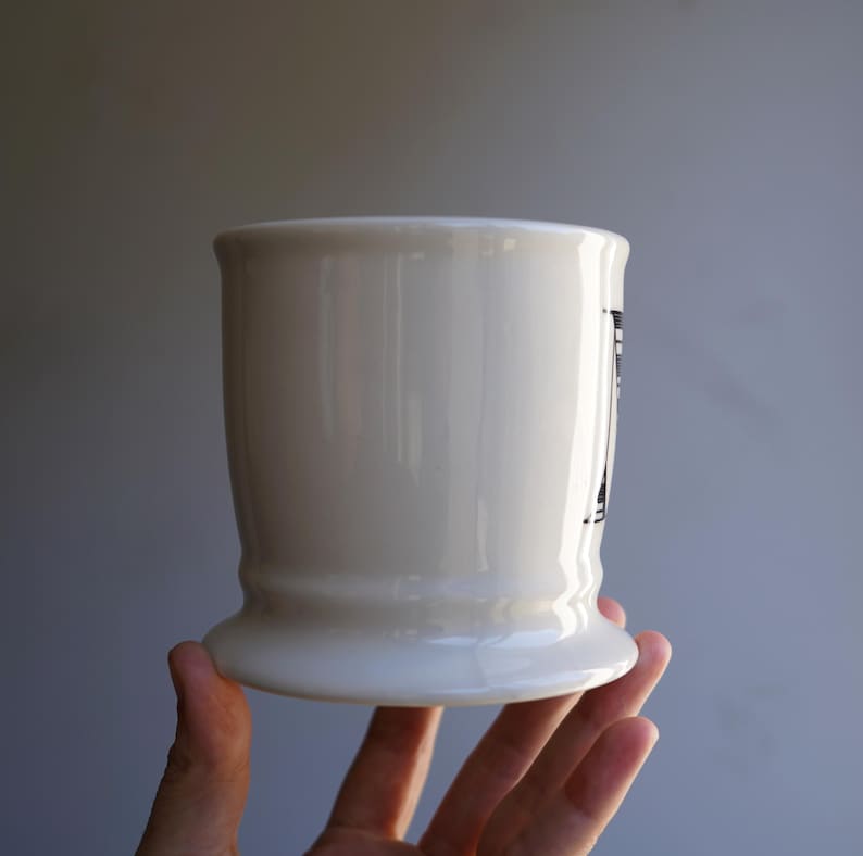 Pu&ograve; includere: Tazza in ceramica bianca con un design bianco e nero. La tazza ha una base larga e un corpo arrotondato.