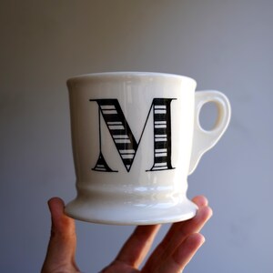 Pu&ograve; includere: Tazza in ceramica bianca con una lettera "M" a strisce bianche e nere sul davanti. La tazza ha una base larga e un manico.