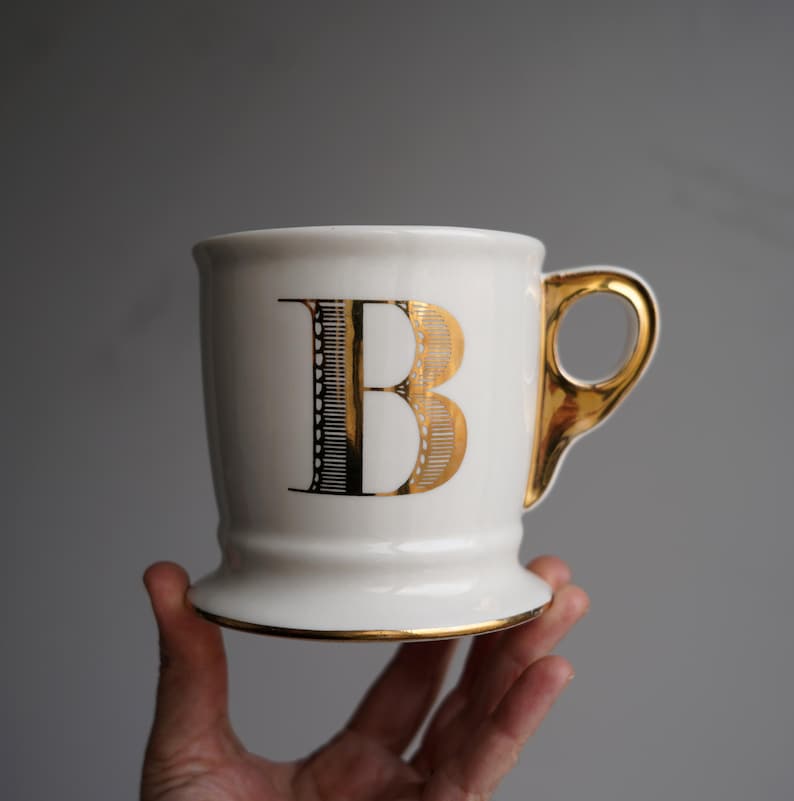 Pu&ograve; includere: Tazza in ceramica bianca con bordo e manico dorati. La tazza ha una lettera "B" dorata sul davanti.