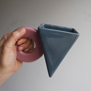 May include: A contemporary ceramic vase with a geometric design. It has a dusty rose circular handle and a triangular, slate blue body. The vase is held in a hand against a neutral background.