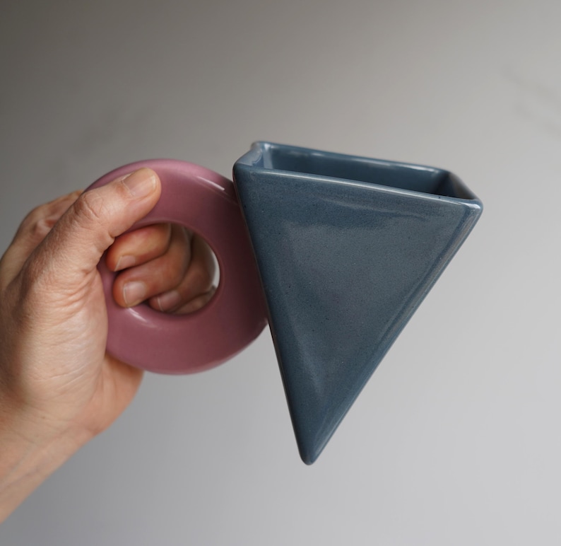 May include: A contemporary ceramic vase with a geometric design. It has a dusty rose circular handle and a triangular, slate blue body. The vase is held in a hand against a neutral background.
