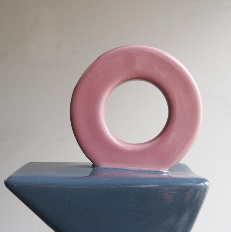 May include: A contemporary ceramic sculpture with a pink circular element on a blue geometric base. The sculpture has a glossy finish and a minimalist design, suitable for home decor.