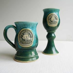 May include: Two ceramic mugs with a teal and green glaze. One is a traditional mug with a handle, and the other is a stemmed goblet. Both feature a circular emblem with text that reads "Maryland Renaissance Festival".