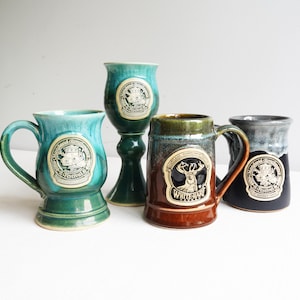 May include: Four ceramic mugs and a goblet in shades of teal, brown, and black. Each piece features a circular emblem with text and a design. One mug has a deer emblem and the text "White Hart".