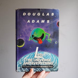 May include: A vintage paperback book titled "Life, The Universe and Everything" by Douglas Adams. The cover features a green cartoon alien with a red tongue against a purple space background with planets and the book's title.