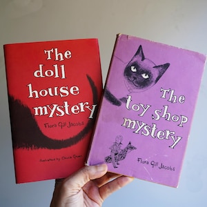 May include: Two hardcover books by Flora Gill Jacobs. The red book is titled The Doll House Mystery and features a black graphic. The purple book is titled The Toy Shop Mystery and features a black cat illustration and a small figure on a horse.