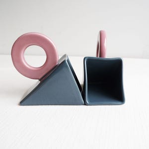 May include: Abstract ceramic bookends in a modern design. The bookends feature a geometric shape with a dusty blue base and a pink ring and handle. The bookends are a decorative item for a bookshelf or desk.