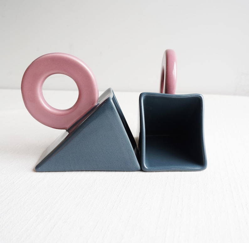 May include: Abstract ceramic bookends in a modern design. The bookends feature a geometric shape with a dusty blue base and a pink ring and handle. The bookends are a decorative item for a bookshelf or desk.