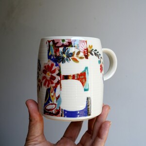 Pu&ograve; includere: Tazza in ceramica bianca con un design floreale colorato con la lettera "E".