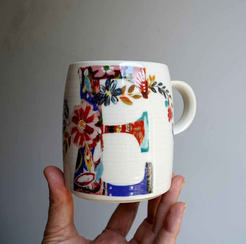 Pu&ograve; includere: Tazza in ceramica bianca con un design floreale colorato con la lettera "E".