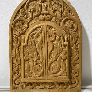 Könnte beinhalten: Ein geschnitztes Wandbehang aus Holz in Bogenform. Das Naturholz weist ein zentrales Doppeltürdesign auf, das von detaillierten floralen und scrollartigen Mustern umgeben ist. Das Stück hat eine hellbraune Farbe.