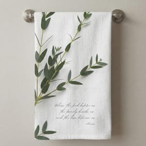 Op de afbeelding: Witte keukendoek met een takje groene eucalyptusbladeren en de tekst "Zegen het voedsel voor ons, de familie naast ons en de liefde tussen ons. Amen." De handdoek hangt aan een zilverkleurige stang.