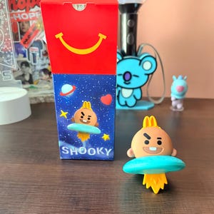 May include: A rectangular cardboard box with a red top featuring a yellow smile and a blue space-themed base with the text SHOOKY. Beside it stands a small brown Shooky character figure with a yellow topknot, riding a teal flying saucer with flames.