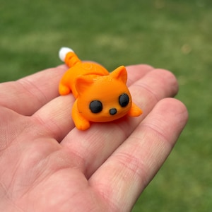 May include: A small, orange cat figurine with black eyes and a white-tipped tail. The cat is lying down. The figurine is held in a hand, with a green background.