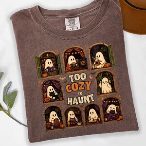 Embrace autumn vibes with this cute "Too Cozy To Haunt" Halloween t-shirt! Featuring adorable ghosts doing cozy indoor activities—reading, baking, drinking hot cocoa, knitting, and listening to music—framed in vintage arched windows. Printed on a comfortable, vintage espresso brown garment-dyed shirt, it’s the ultimate spooky season apparel for book lovers, Halloween enthusiasts, and fans of aesthetic fall fashion. Perfect casual Autumn gift!