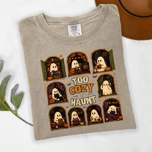 Puede incluir: Una camiseta beige de cuello redondo con un estampado gr&aacute;fico de ocho ventanas arqueadas con fantasmas realizando actividades acogedoras. El texto central dice "TOO COZY TO HAUNT" en letras naranjas y blancas. Etiqueta Comfort Colors.