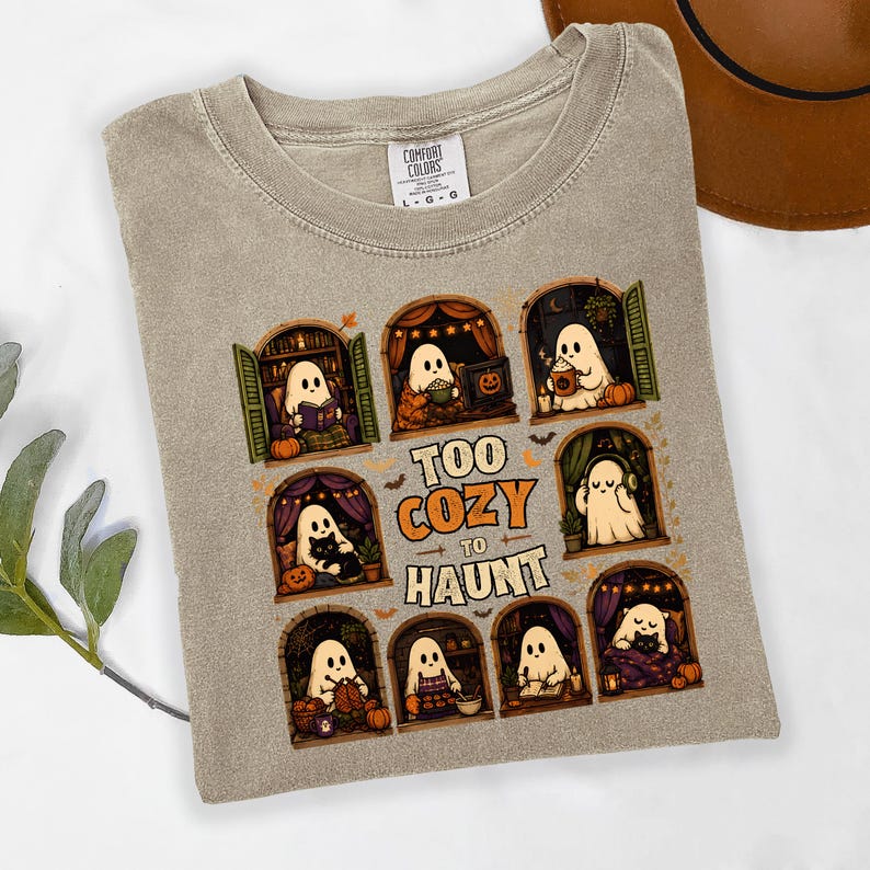 Puede incluir: Una camiseta beige de cuello redondo con un estampado gr&aacute;fico de ocho ventanas arqueadas con fantasmas realizando actividades acogedoras. El texto central dice "TOO COZY TO HAUNT" en letras naranjas y blancas. Etiqueta Comfort Colors.