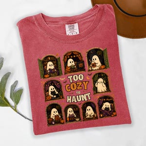Puede incluir: Una camiseta roja deste&ntilde;ida con un estampado gr&aacute;fico de ocho ventanas arqueadas, cada una con un fantasma blanco realizando actividades acogedoras como leer, beber y tejer. El texto "TOO COZY TO HAUNT" est&aacute; centrado en letras naranjas y blancas.