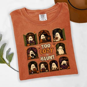 Puede incluir: Una camiseta de color naranja quemado con un estampado gr&aacute;fico de ocho ventanas arqueadas, cada una con un fantasma blanco realizando actividades acogedoras. El texto central dice "TOO COZY TO HAUNT" en una fuente con borde naranja. Cuello redondo.