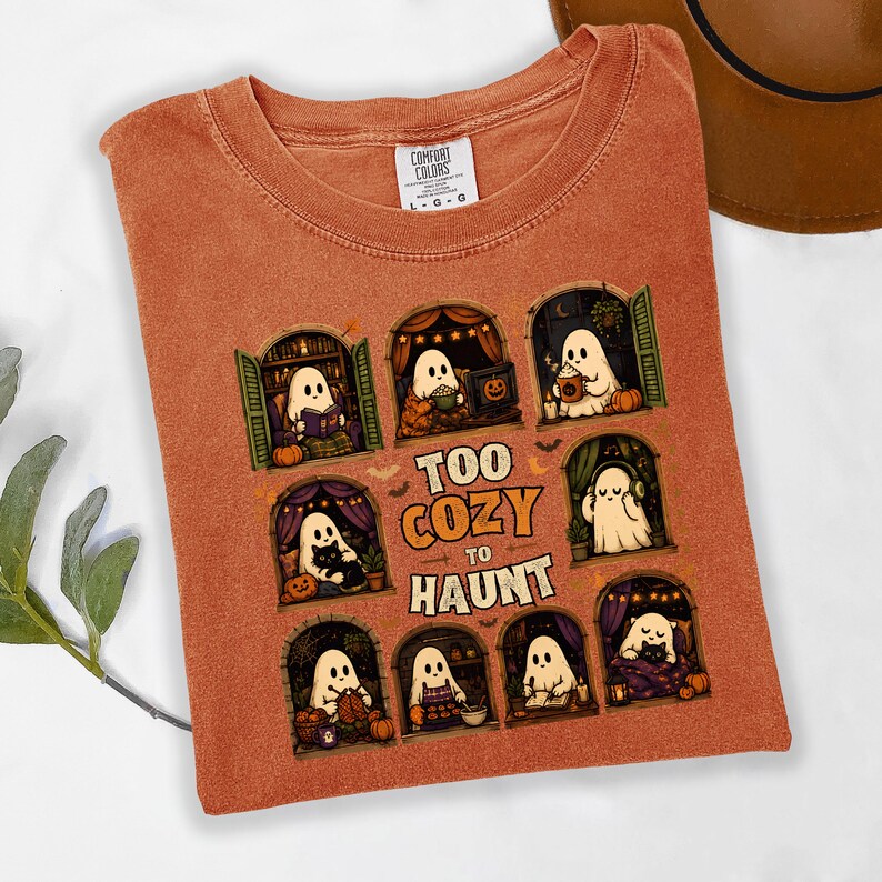 Puede incluir: Una camiseta de color naranja quemado con un estampado gr&aacute;fico de ocho ventanas arqueadas, cada una con un fantasma blanco realizando actividades acogedoras. El texto central dice "TOO COZY TO HAUNT" en una fuente con borde naranja. Cuello redondo.