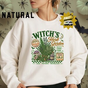 A vintage botanical illustration on a natural cream sweatshirt. Bold green “WITCH’S” sits above orange script “Herb Garden,” flanked by a crescent moon and stars. At the center, a tied bundle of lavender and leafy herbs is surrounded by purple mushrooms, wildflowers, and a “WITCHES BREW” bottle. Circular stamps read “FRESHLY GATHERED,” “LOCALLY GROWN HERBS,” and “MOONLIT GROWN · AUTUMN PICKED.” A green-and-white checkered banner at the bottom states.
