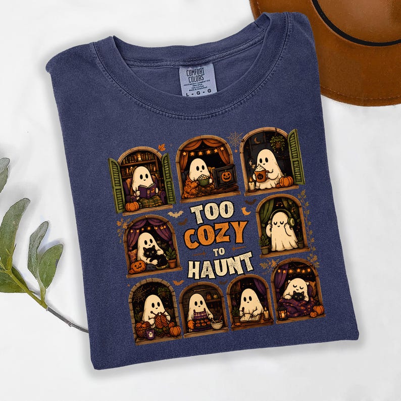 Puede incluir: Una camiseta azul marino con un estampado gr&aacute;fico de ocho ventanas arqueadas, cada una con un fantasma blanco realizando actividades acogedoras. El texto central dice "TOO COZY TO HAUNT" en letras de color naranja y blanco.