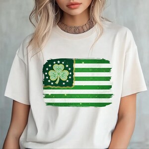 以下が含まれることがあります： クリーム色のTシャツ。緑と白の旗のデザインで、クローバーと星が描かれています。旗のデザインは、ブラシストロークの効果があります。このシャツは、カジュアルな普段着です。