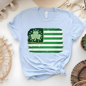 以下が含まれることがあります： 緑と白の旗のデザインが特徴のライトブルーのTシャツ。クローバーと星が描かれています。旗は、使い古された筆致効果があります。Tシャツは柔らかい素材で作られています。