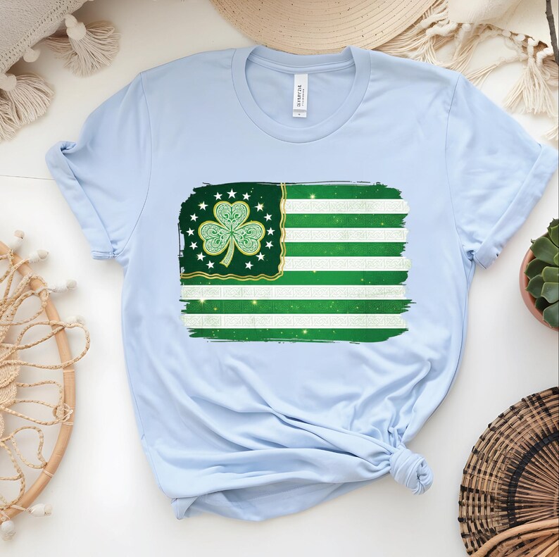 以下が含まれることがあります： 緑と白の旗のデザインが特徴のライトブルーのTシャツ。クローバーと星が描かれています。旗は、使い古された筆致効果があります。Tシャツは柔らかい素材で作られています。
