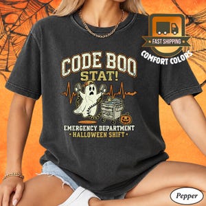 May include: A charcoal gray short-sleeve t-shirt featuring a graphic of a ghost wearing a baseball cap and holding defibrillator paddles. The text reads CODE BOO STAT! EMERGENCY DEPARTMENT HALLOWEEN SHIFT, accompanied by a medical cart and pumpkin.