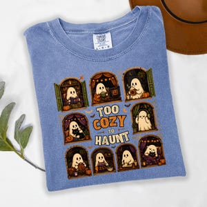 Puede incluir: Una camiseta azul con un gr&aacute;fico de fantasmas en varios entornos acogedores y el texto "TOO COZY TO HAUNT". El dise&ntilde;o incluye fantasmas leyendo, bebiendo y relaj&aacute;ndose en ventanas arqueadas, rodeados de peque&ntilde;as calabazas y murci&eacute;lagos.