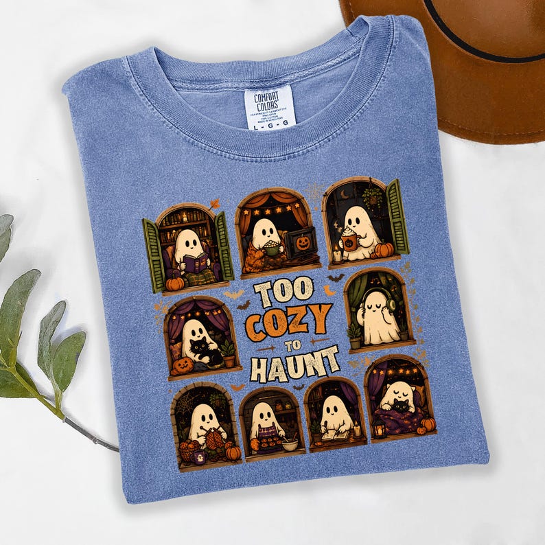 Puede incluir: Una camiseta azul con un gr&aacute;fico de fantasmas en varios entornos acogedores y el texto "TOO COZY TO HAUNT". El dise&ntilde;o incluye fantasmas leyendo, bebiendo y relaj&aacute;ndose en ventanas arqueadas, rodeados de peque&ntilde;as calabazas y murci&eacute;lagos.