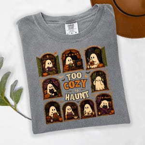 Puede incluir: Una camiseta gris con un estampado gr&aacute;fico de ocho ventanas arqueadas, cada una con un fantasma de dibujos animados realizando actividades acogedoras como leer, tejer y beber caf&eacute;, con el texto "TOO COZY TO HAUNT" centrado en el medio.