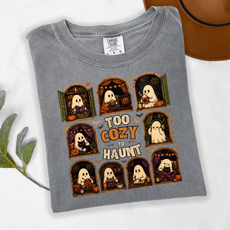 Puede incluir: Una camiseta gris con un estampado gr&aacute;fico de ocho ventanas arqueadas, cada una con un fantasma de dibujos animados realizando actividades acogedoras como leer, tejer y beber caf&eacute;, con el texto "TOO COZY TO HAUNT" centrado en el medio.