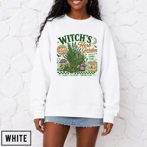 May include: A white long-sleeve sweatshirt featuring a graphic print with the text Witch's Herb Garden, Freshly Gathered, Locally Grown Herbs, Moonlit Grown, and Autumn Picked, alongside illustrations of herbs, mushrooms, and a small potion bottle.