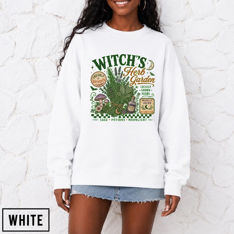 May include: A white long-sleeve sweatshirt featuring a graphic print with the text Witch's Herb Garden, Freshly Gathered, Locally Grown Herbs, Moonlit Grown, and Autumn Picked, alongside illustrations of herbs, mushrooms, and a small potion bottle.