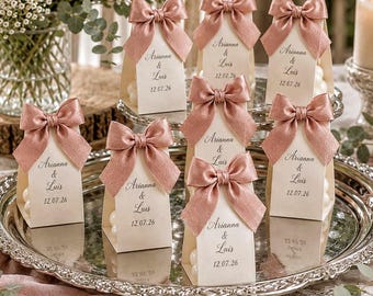May include: A collection of wedding favor boxes with pink satin bows, arranged on a silver platter. Each box has a tag with the names "Arianna & Luis" and the date "12.07.26". The scene includes flowers and a lit candle.